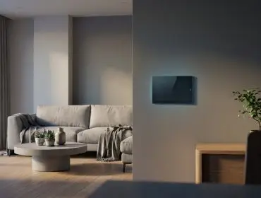 Écran de contrôle tactile fixé au mur d'un salon élégant et lumineux avec un canapé gris et une table basse.